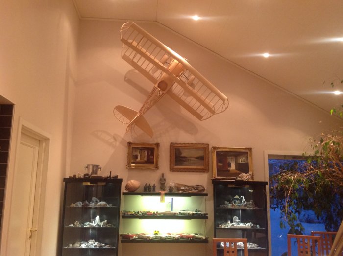 Modellflygplan upphängt på en hög innervägg omgiven av konst och skåp med samlarföremål.