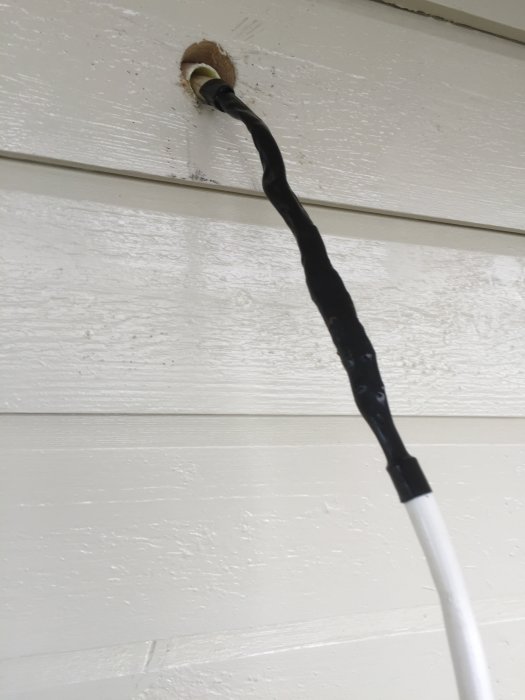 Elkabel med extra krympslang genom vägguttag på en vitmålad trävägg, installationsdetalj för balkongbelysning.