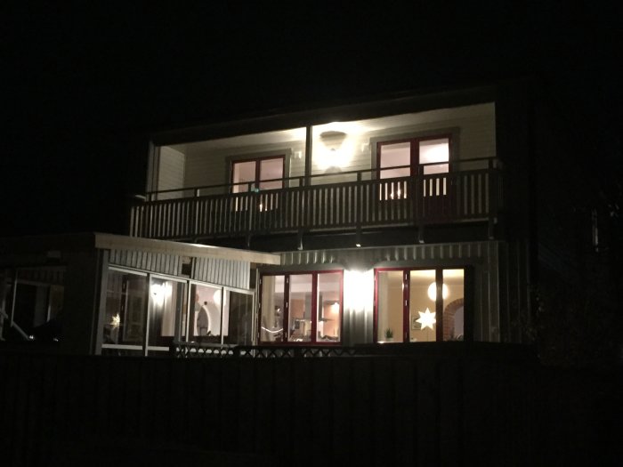 Kvällsbild av ett hus med tända lampor, nya balkonglampan synlig, i höstmörkret.