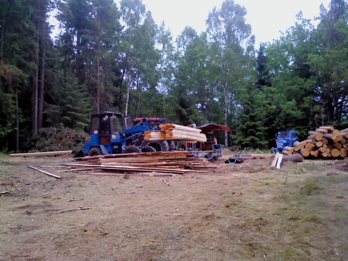 En byggplats i skogsmiljö med en hög av bondsågade träplankor och stockar bredvid en blå traktor och en liten grön grävmaskin.