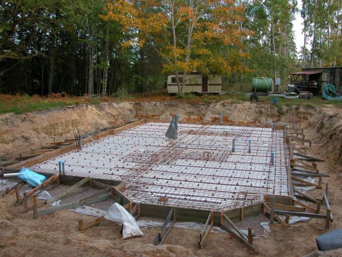 Grund till hus med armeringsjärn i skogsmiljö, grävmaskin och byggmaterial i bakgrunden.