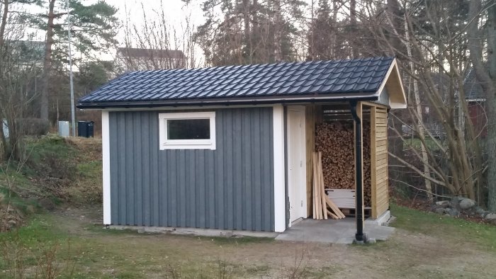 Ett nyligen uppfört förråd med grå panel och svart takplåt, nästan färdigt med vedstapel och byggmaterial synligt.