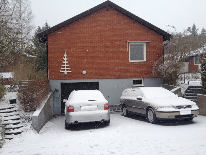 Vinterbild med snötäckta bilar framför hus med julgran av ljusslingor på väggen.