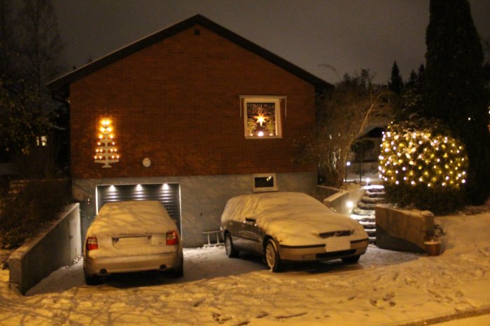 Snöbelagda bilar framför hus med juldekorationer och upplyst gran i kvällsmörkret.