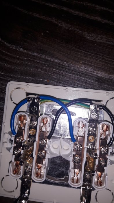 Öppen 4-vägs stickkontakt med blå och svart kabel fastskruvade, gulgrön kabel löst liggande ovanpå mot mörkt underlag.