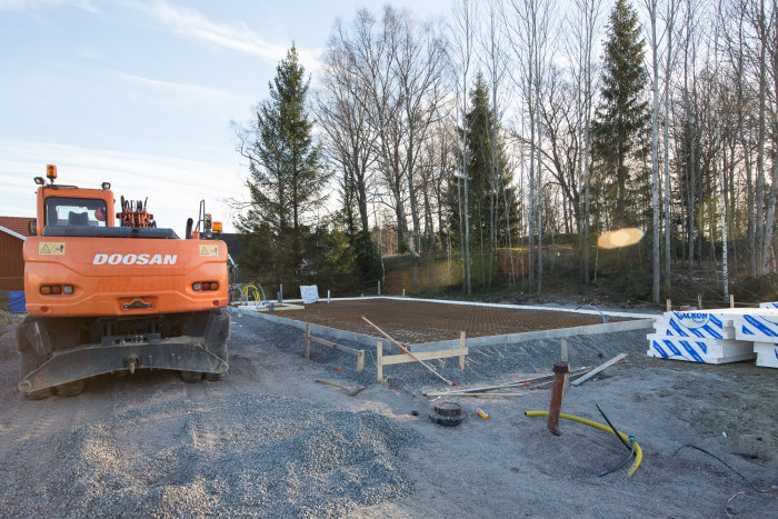 Armerad betongplatta på mark förberedd för gjutning, med byggmaskin och skog i bakgrunden.