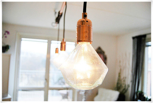 Modern taklampa med industriell design och exponerad ljuskälla, upphängd i ett rum med suddig bakgrund.