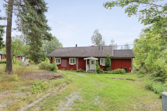 Rött trähus med vita knutar omgivet av träd och grön vegetation, dagtid, molnigt, svensk sommarstuga.