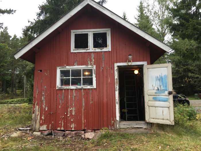 Förslitet rött trähus med flagnande färg och en delvis öppen dörr som avslöjar en rörig inredning, omgivet av träd och vegetation.