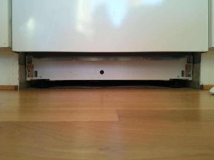 Bild på undersidan av en vit maskin med borttaget skydd som avslöjar inre delar och golvkontakt.