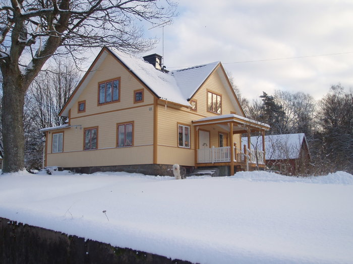 Ett tvåvåningshus med ljusgul fasad och snötäckt tak omgivet av snö och träd.