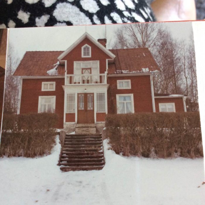 Ett rött hus med vita fönsterkarmar och balkong, fotograferat 1965, omgivet av snö och vinterlandskap.