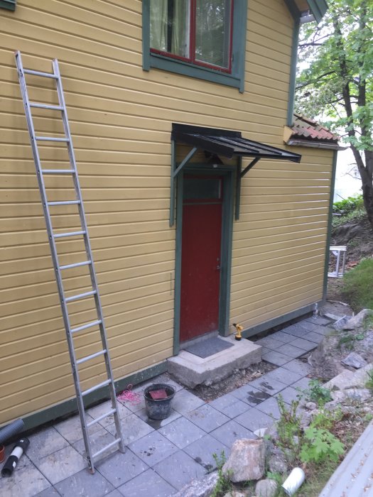 Sandgul träfasad med oxidgröna foder kring fönster och oxidröd dörr på en tvåvåningshus med trädgårdsstege vid sidan.