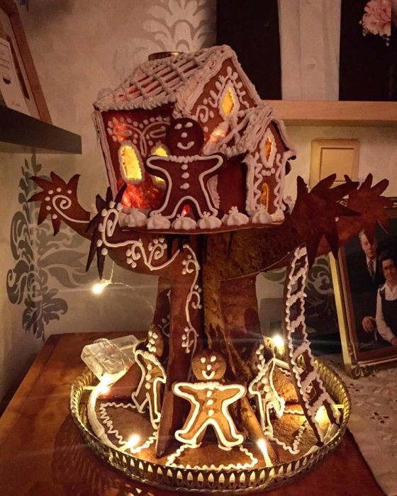 Ett hemmagjort pepparkakshus formad som ett trädhus med upplysta fönster och pepparkaksfigurer runt omkring, placerat på ett fat.