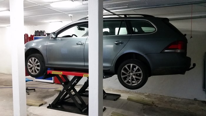 En bil upplyft på en svart billyft i ett garage med betonggolv och vitmålade väggar.