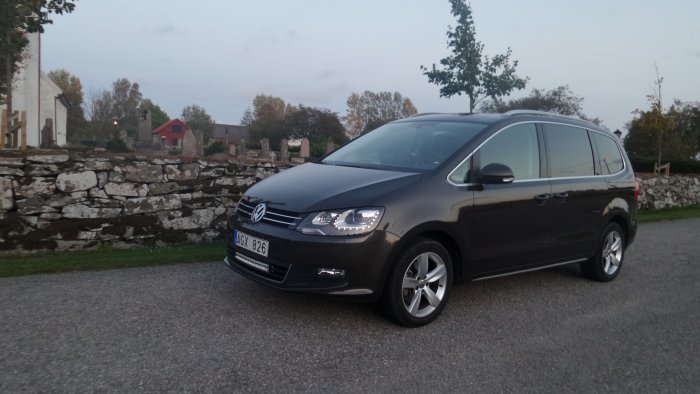 Volkswagen Sharan parkerad framför stenmur med tända strålkastare.