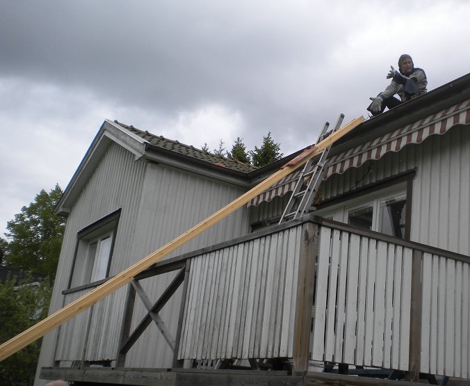 Person på tak till grått hus som skickar ner en bräda längs med en skyffel.