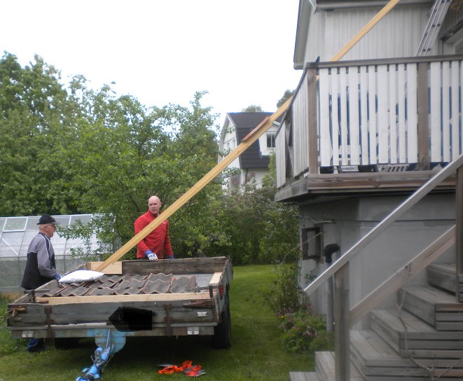 Två personer lastar avbrutna träplankor från en släpvagn nära ett hus med trädgård.