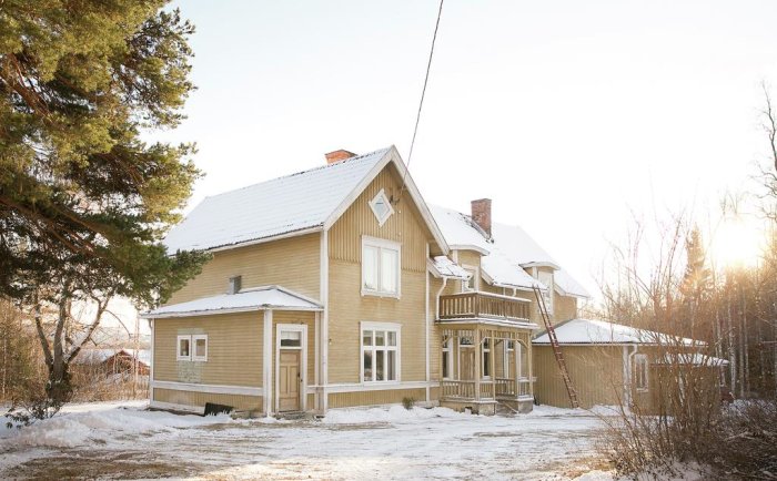 Välbevarat gulmålat trähus med snötäckt tak och balkong i vintersol, omgivet av snö och träd.