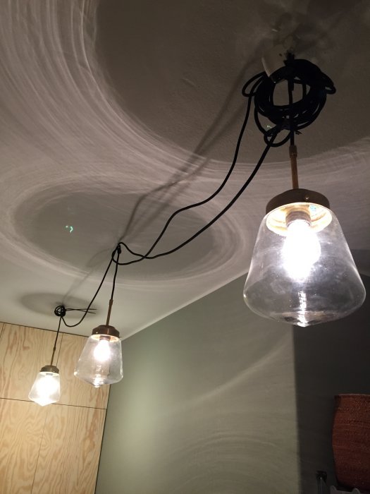 Tre upphängda lamparmaturer med synliga svarta textilkablar som löper mellan lampor och en takkrok.