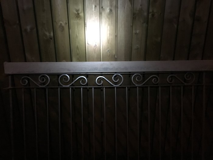 Svetsat metallstaket med dekorativa krön mot trävägg i dålig belysning.