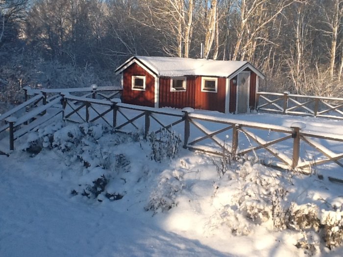 Röd stuga med vita knutar i vinterlandskap med snötäckt staket och träd i bakgrunden.