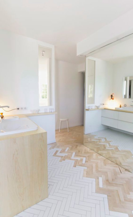 Ljus parkettgolv möter vitt kakel i en herringbone-mönster i minimalistiskt badrum.