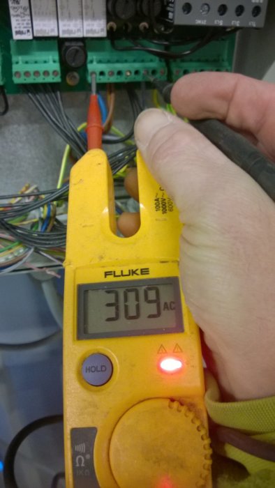 Ett digitalt multimeter i förgrunden visar 309 volt AC under en mätning av elektriska kablar i en elkoppling.