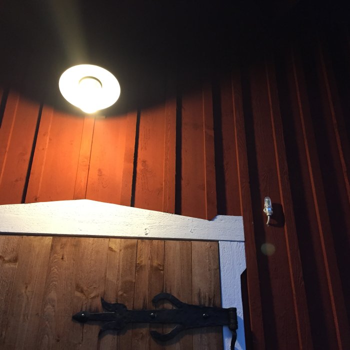 Tänd lampa på trävägg, skymningsrelä monterat till höger om lampan nära en dörr med svart gångjärn.