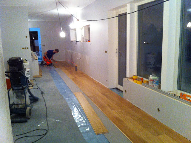 Läggning av ljusa trägolvplank i en inomhusmiljö med en person i arbete och byggmaterial.