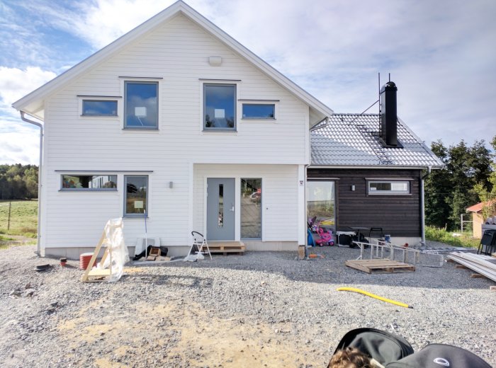 Vit tvåvåningshus med mörk tillbyggnad och byggmaterial framför, under en klar himmel.