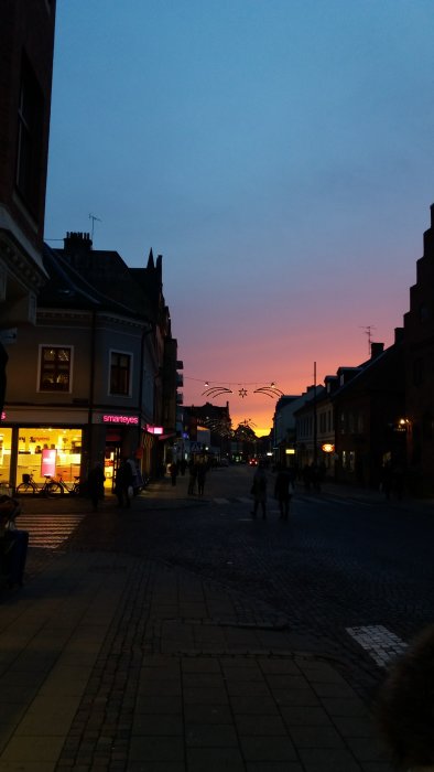 Stadsgata vid skymning med människor och butiker, färgglad himmel i bakgrunden.