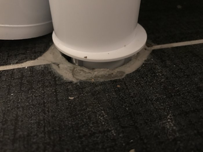 Vit avloppsröranslutning till handfat utan synlig tätning, omgiven av klinkergolv och silikonrester.