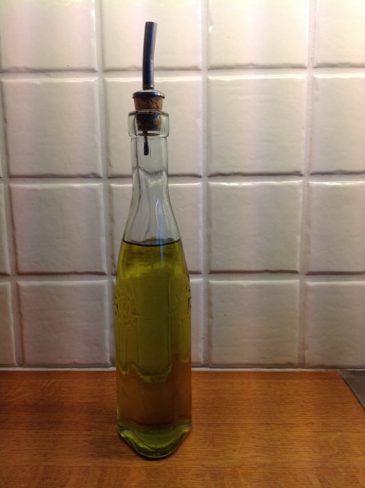 Glasflaska med olivolja och en kork med pip mot en kaklad väggbakgrund.