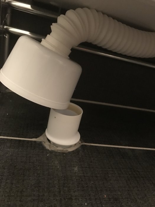 Vit avloppsrörskoppling under vask går genom svart klinkergolv med synliga silikonrester och utstickande små pluggar.