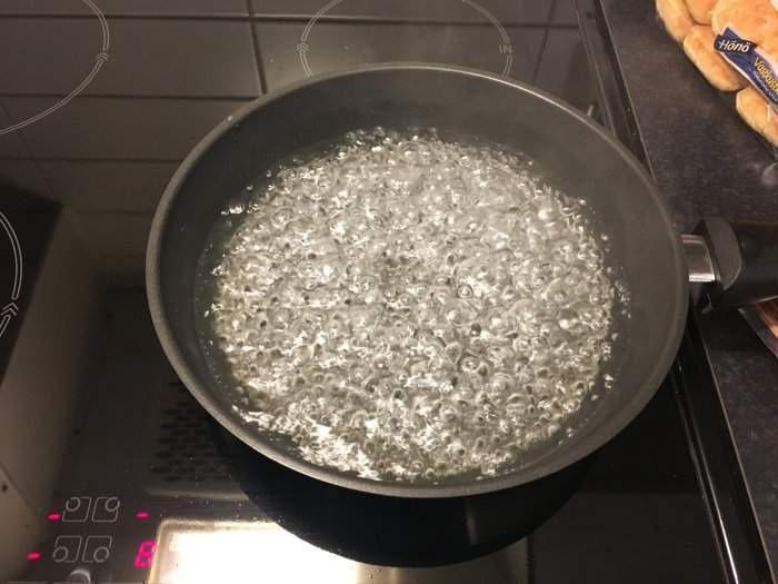 En stekpanna på en induktionshäll full med bubbelande vatten vid kokpunkt, omgiven av köksutrustning.