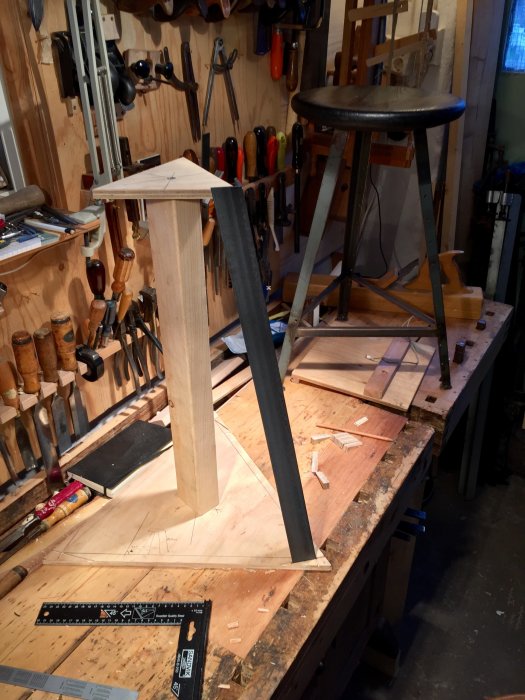 Arbetsbänk i snickeri med pågående träprojekt, omgiven av verktygsvägg och pall.