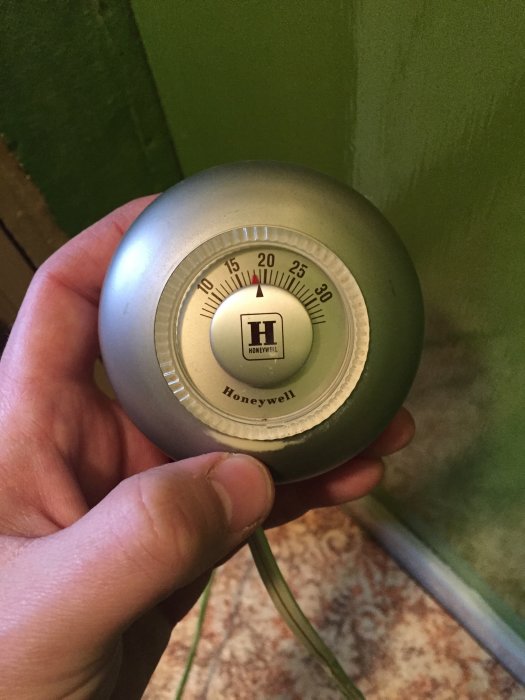 Hand håller gammal rund Honeywell-termostat med synliga inställningsskalor mot en grön vägg.