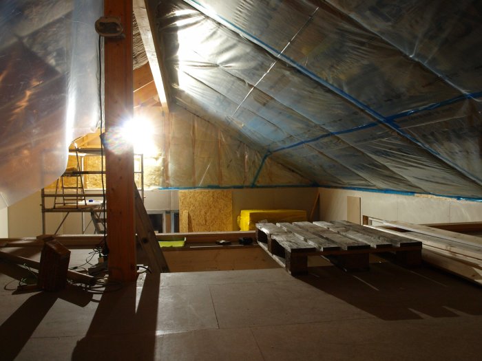 Isolerat vindutrymme med plasttäckt tak och trästomme, isolermaterial och byggmaterial syns.