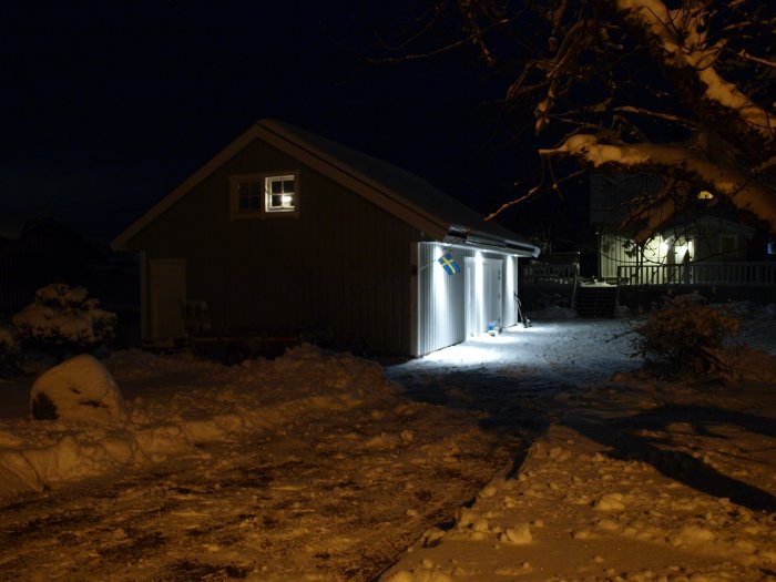 Upplyst friggebod på kvällen med snö runtomkring och en svensk flagga på sidan.