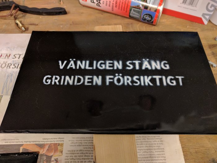 Svart skylt med texten "VÄNLIGEN STÄNG GRINDEN FÖRSIKTIGT" applicerad med vit sprayfärg och schablon.