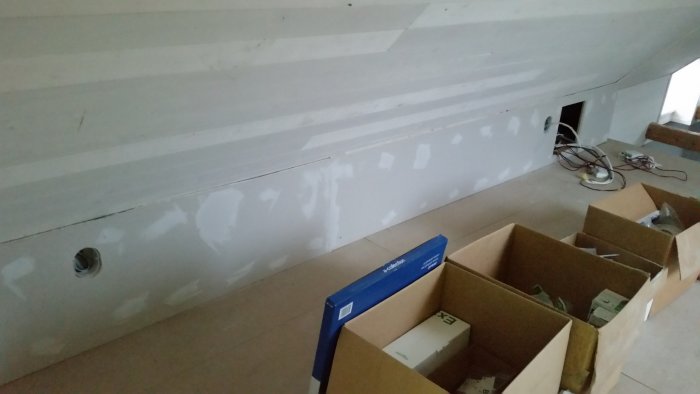 Vägg med spackling och slipspår, elutrustning och kartonger på golv i teknikrum under renovering.