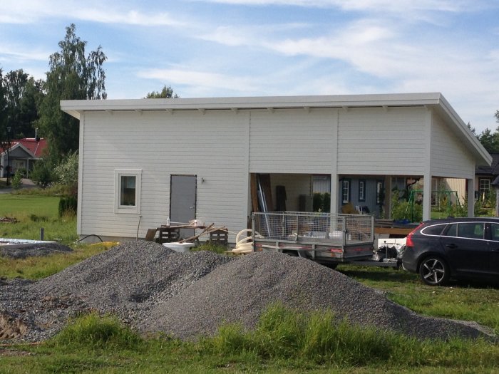 Nybyggd carport med förråd/gäststuga, omgiven av byggmaterial och grushög på en plan tomt.