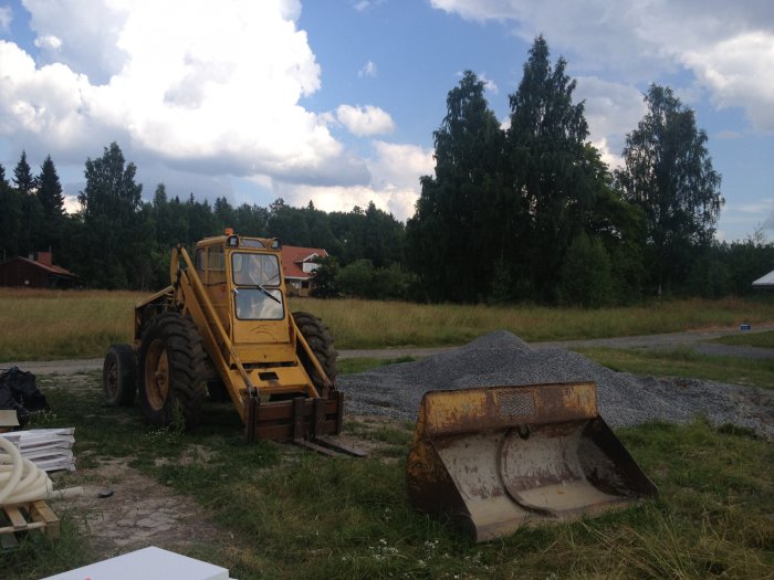 Gul baklastare vid en hög med grus på en byggarbetsplats med träd i bakgrunden.