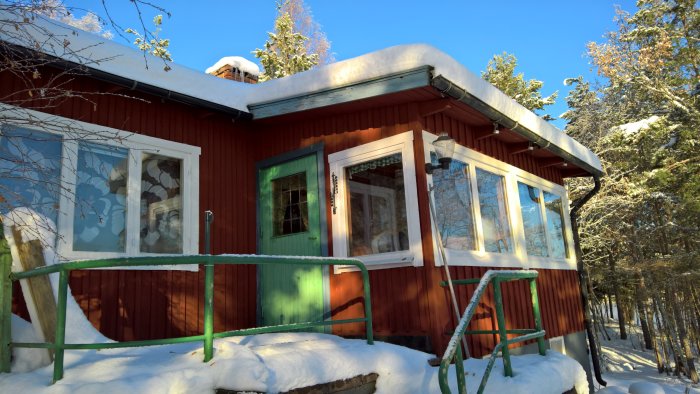 Röd stuga med tillbyggnad och stora teakfönster, omgiven av snö och skog i soligt väder.