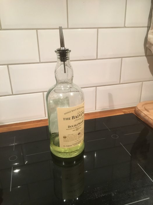 Glasflaska med etikett och hällpip står på svart spis framför vit kakelvägg.
