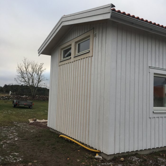 Hörnet av ett nybyggt vitt hus med två fönster och plåtfasad, gul gasledning längs marken.