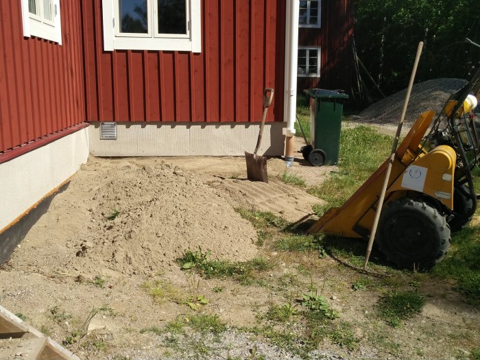 Pågående arbete på uteplats med grävmaskin, spadar och sand framför röd träfasad.