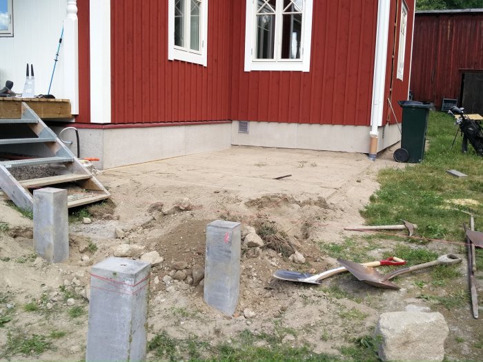 Pågående arbete på en uteplats med nedgrävda plintar och byggmaterial framför ett rödmålat hus.