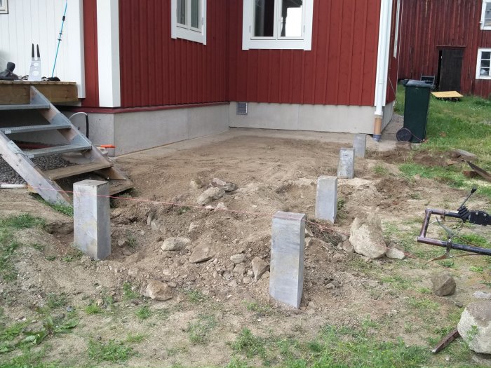 Pågående anläggning av uteplats med nedgrävda betongplintar nära röd husvägg, singel, verktyg och grävmaskin synlig.